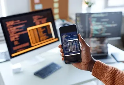 Hand holding a smartphone that shows code, in front of computer monitors displaying source code