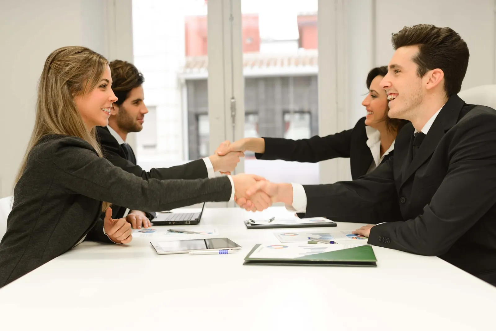 Two business teams smiling and shaking hands across a meeting table with documents and a laptop